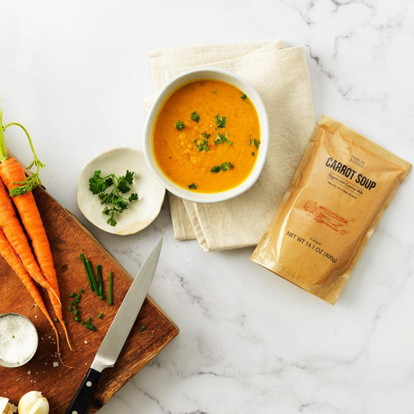 cutting board with carrots, sharp cutting knife, salt, green herbs, bowl of creamy carrot soup, bag of public goods carrot soup