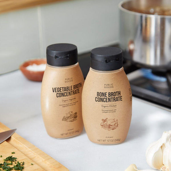 bottle of organic vegetable bone broth concentrate, bottle of organic chicken bone broth concentrate, cutting board and cutting knife, pot on heated stove, garlic clove, kitchen counter