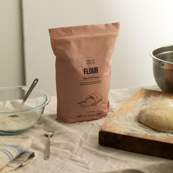 bag of organic all purpose flour, flour in glass bowl, dough on wooden cutting board, metal measuring spoon, kitchen towel, metal mixing bowl