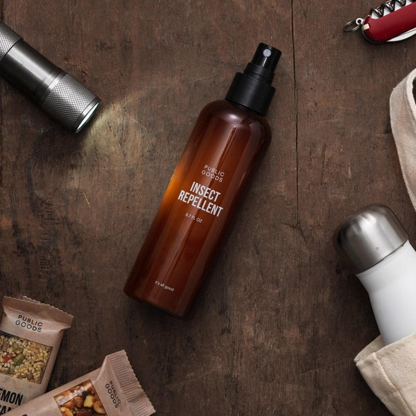 bottle of public goods insect repellent on wooden table, flashlight, water bottle, snack bars, tote bag, wine bottle opener