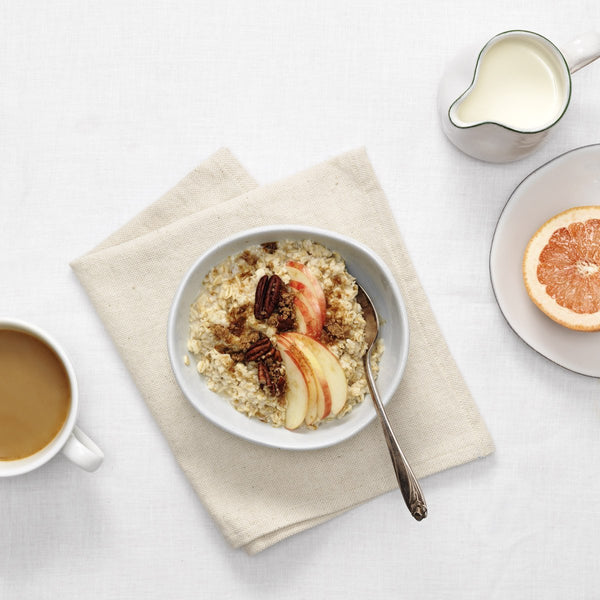 bowl of oatmeal with pecans and sliced apples, halved grapefruit on a plate, cup of coffee, carafe of milk, tablecloth