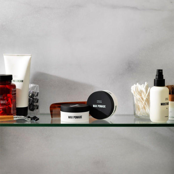 two containers of wax based hair pomade, qtips in glass jair, bottle of moisturizer, mens daily vitamins, shaving razor, razor blades, shaving cream, glass bathroom counter