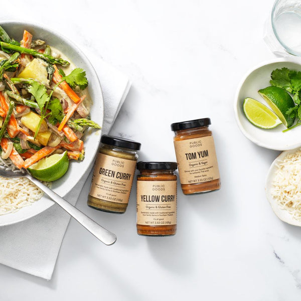 jar of green curry, jar of yellow curry, jar of tom yum curry, rice and curried vegetables on a plate, bowl of rice, spoon, napkin, lime wedges and basil in bowl